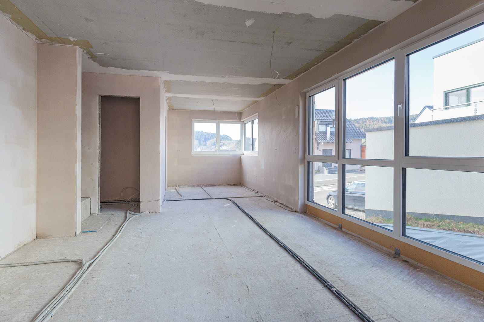 Vorher-Bild für virtuelles Home Staging: Ein heller, langgestreckter Raum im Rohbauzustand. Die Baustelle zeigt einen nackten Betonboden mit freiliegenden Leitungen, unverputzte Wände und eine unfertige Decke. Große, moderne Fensterfronten sorgen für viel Tageslicht und bieten ideales Potenzial für die digitale Visualisierung eines offenen Essbereichs.